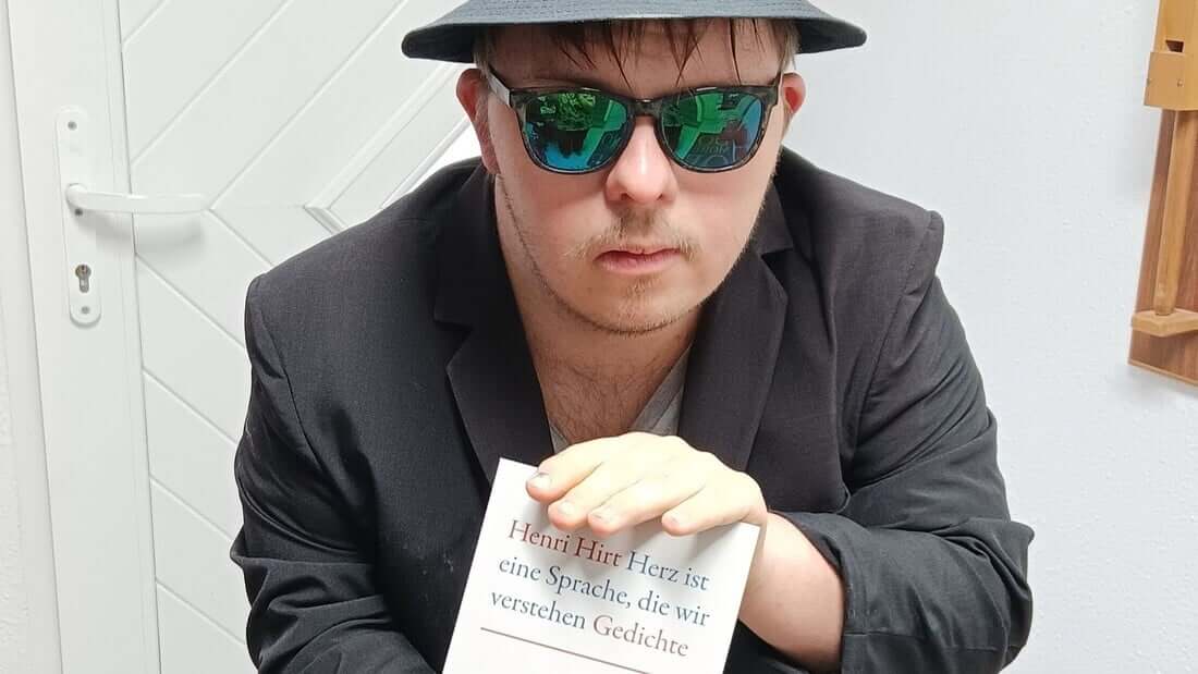 Die ist ein Foto von Henri Hirt. Er trägt einen Hut und eine verspiegelte Sonnenbrille. Er hält sein Buch in die Kamera. Das Buch hat ein weißes Cover und heißt „Herz ist eine Sprache, die wir verstehen”.